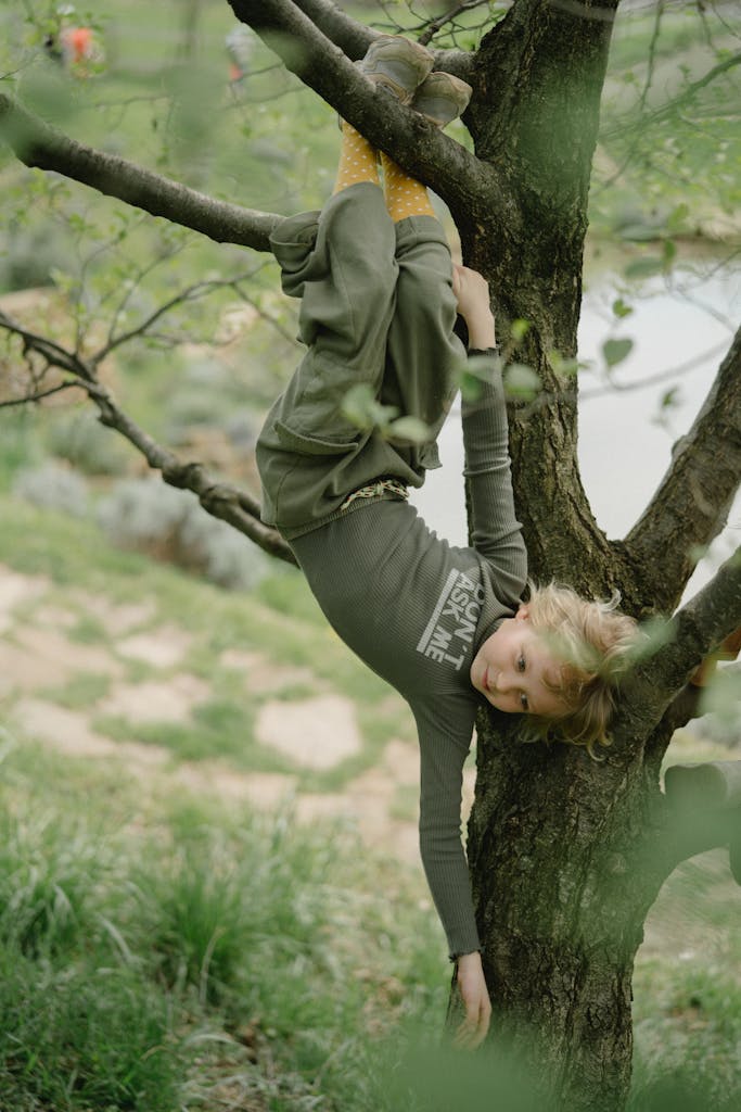 A young child playfully hangs upside down from a tree branch outdoors in a park setting.
