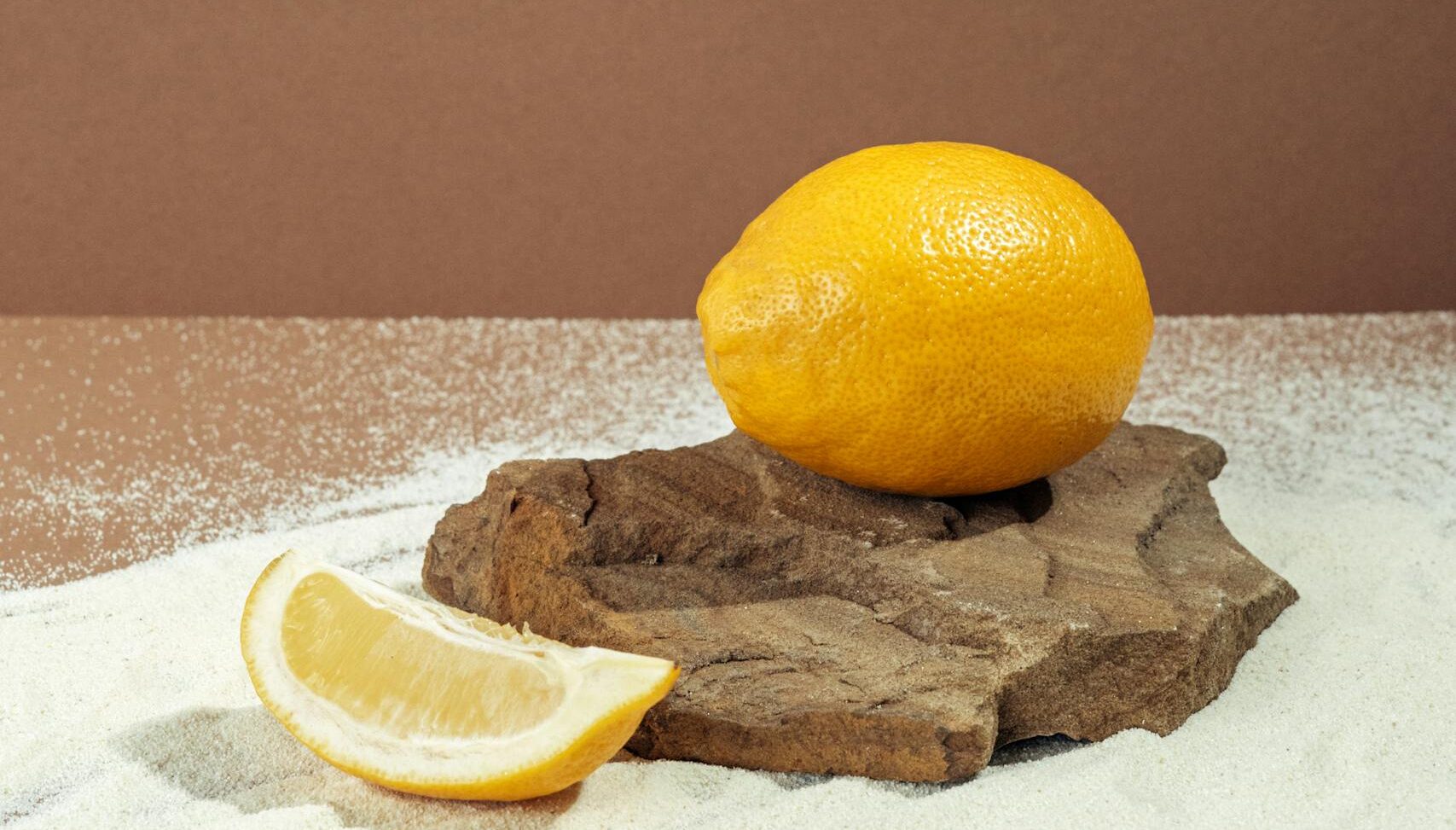 A fresh lemon and slice on a wooden surface with white sand and brown background.